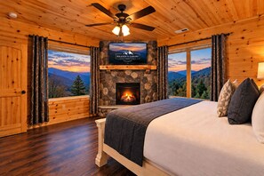 A cozy bedroom with a stone fireplace, a bed, and a large window offering a mountain view.