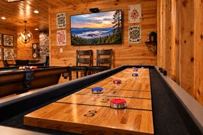 A shuffleboard table with numbered spots and pucks, a television displaying a scenic landscape, and wooden walls with framed playing cards.