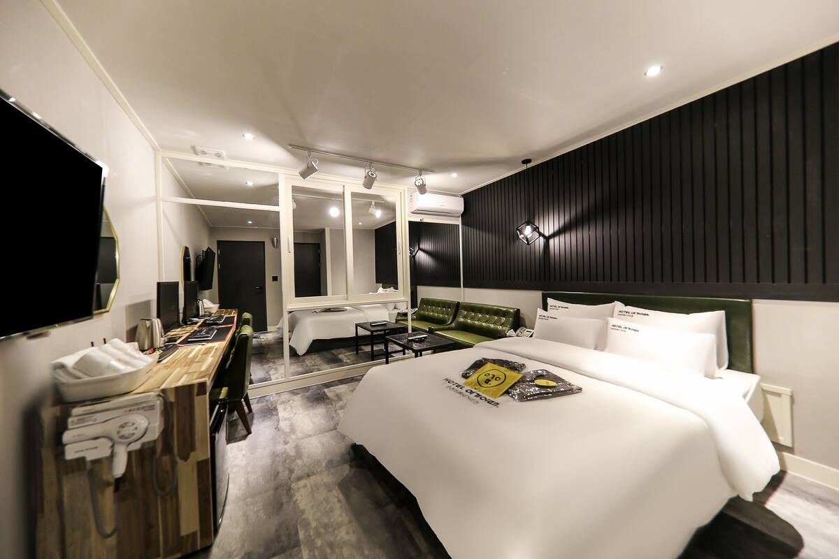 A modern hotel room with a large bed, a desk with a telephone, and a seating area with a green sofa.