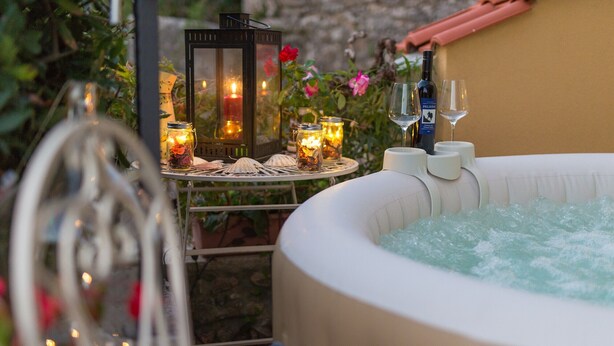 Tinaja de hidromasaje al aire libre con velas y una botella de vino, rodeada de follaje y flores.