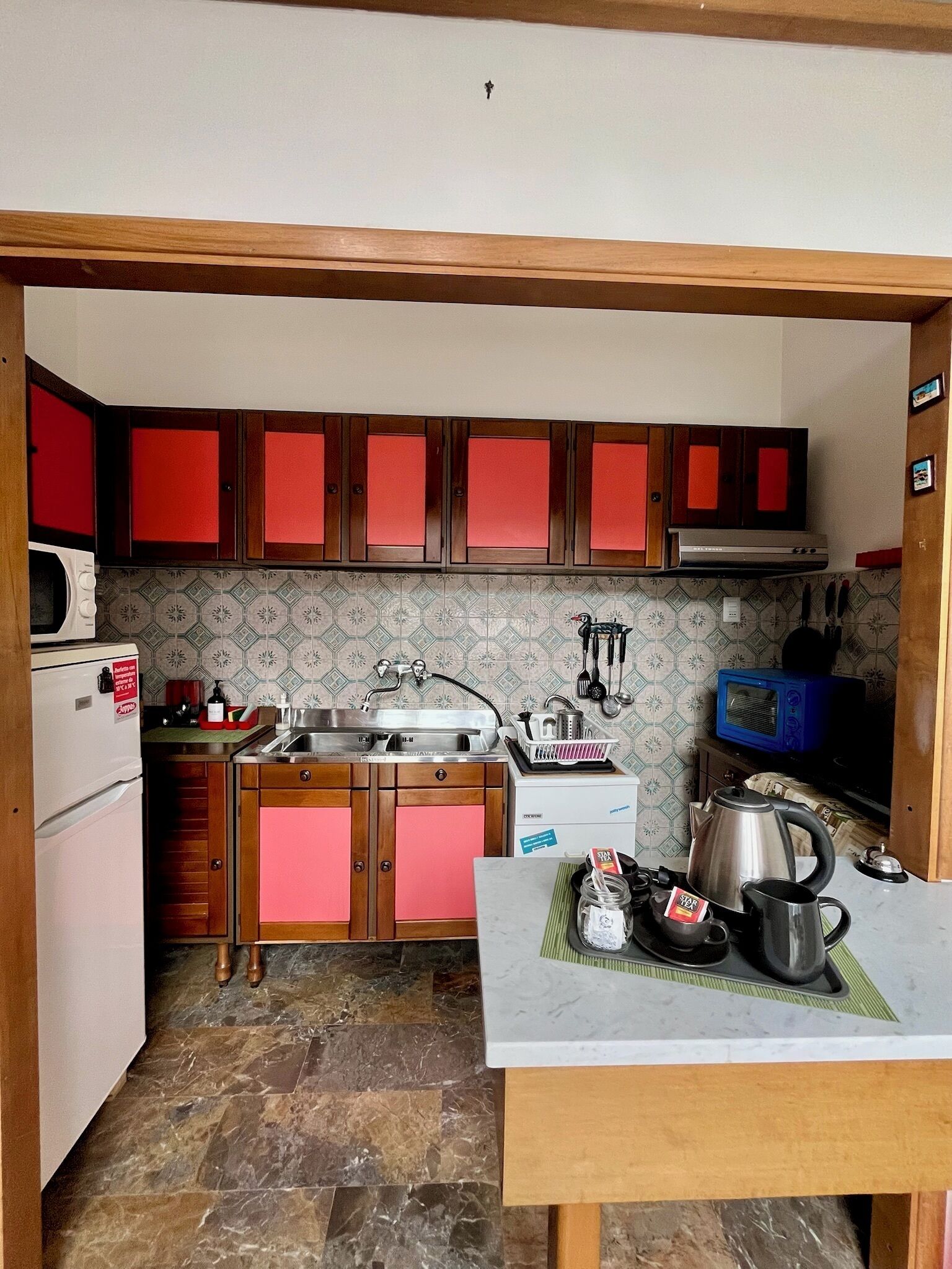 Una cocina compacta con armarios de madera, un lavamanos de acero inoxidable, un microondas y una tetera sobre la encimera.