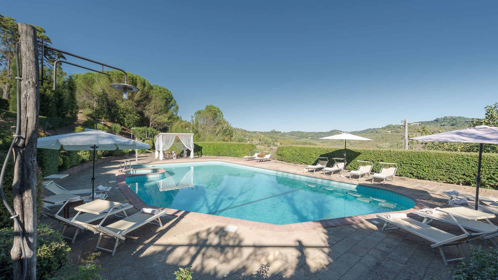 A swimming pool with lounge chairs and umbrellas, surrounded by greenery and hills.