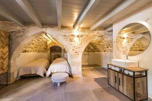A stone-walled bedroom with two beds, a wooden bench, and a large mirror.