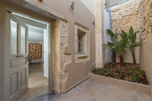 A hallway with a stone wall, a door leading to a room, and a small window. There is a planter with flowers and a palm tree. - Casa Vitae Villas with private pool -  Myrthia (Rethymno)