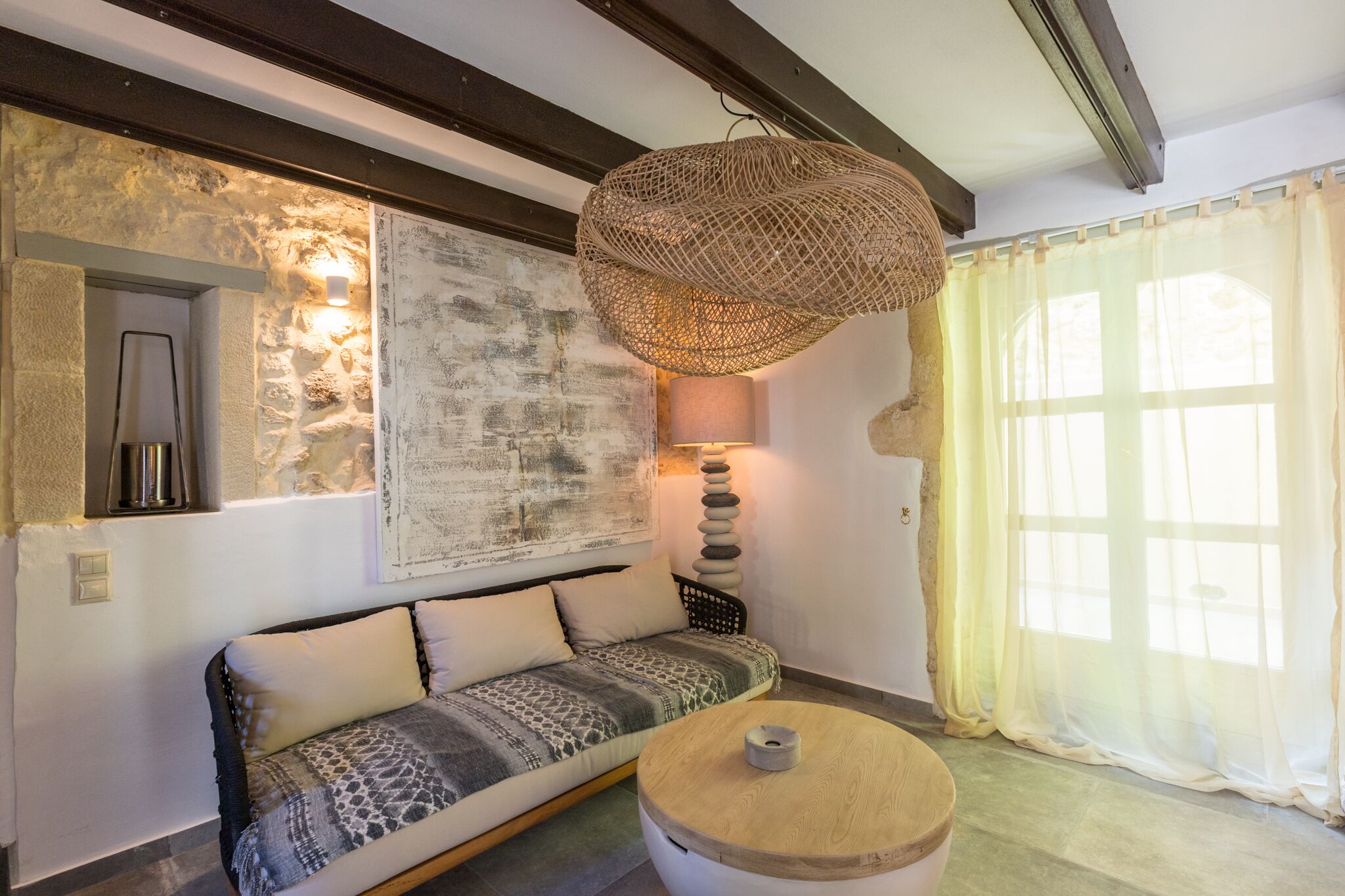 Una sala de estar acogedora con un sofá estampado, una mesa de centro redonda de madera, un colgante de luz tejido y un ventanal con cortinas translúcidas.
