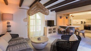 Un salon moderne avec un canapé, une table basse ronde et des fauteuils en osier. Un téléviseur est posé sur un meuble en bois, et on aperçoit une cuisine au fond.