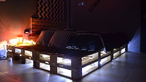 Ein Bett mit Holzrahmen und Matratze auf einer Plattform, eine Lampe links sowie eine Wand mit Zickzackmuster und ein Schild mit japanischen Schriftzeichen.