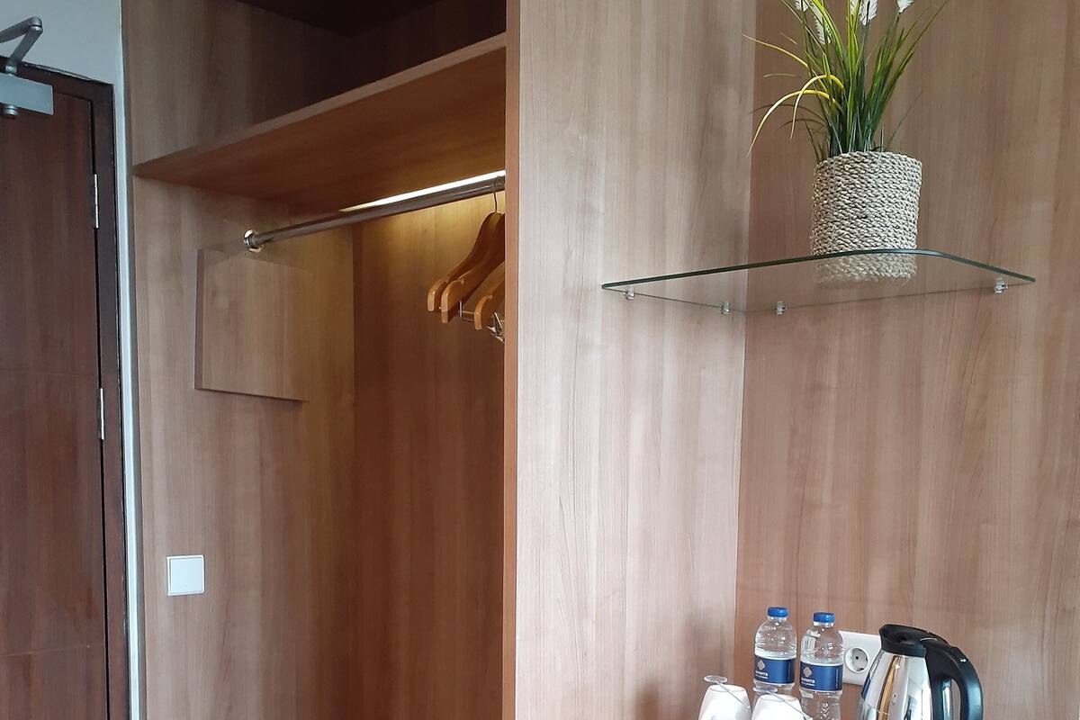 A modern hotel room with a wooden wardrobe, a small seating area with a glass shelf, a compact kitchenette, and a potted plant.