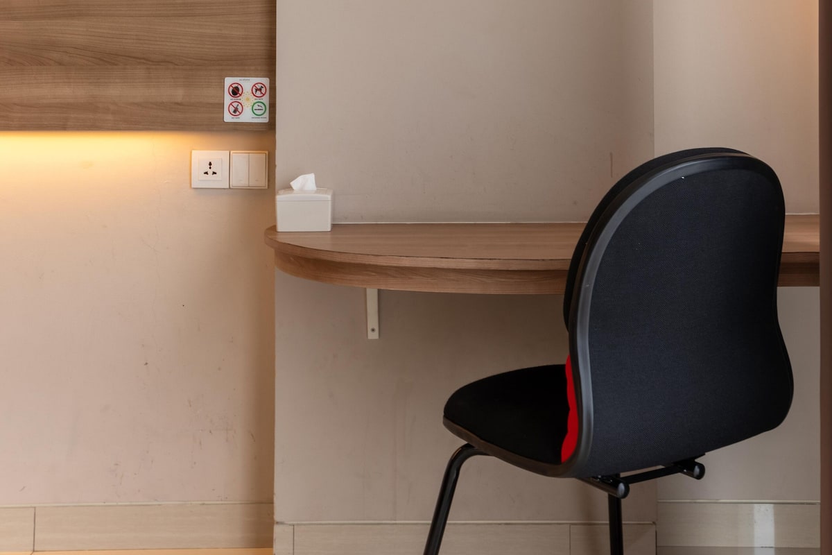 A modern office chair with a black seat and red accents, positioned next to a wooden desk against a wall with a light switch and a no-smoking sign.