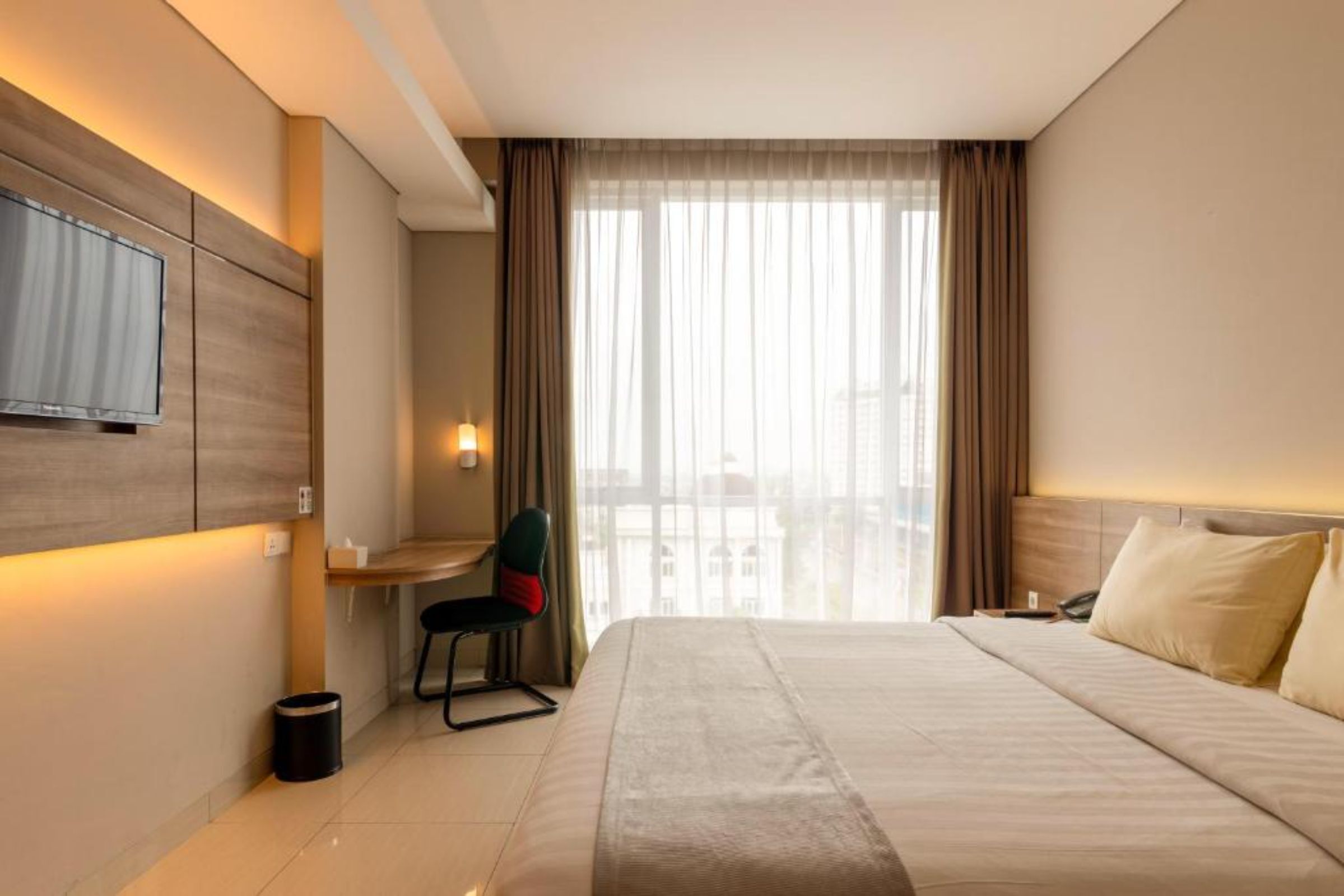 A hotel room with a bed, a desk, a chair, a TV mounted on the wall, and a window with curtains.