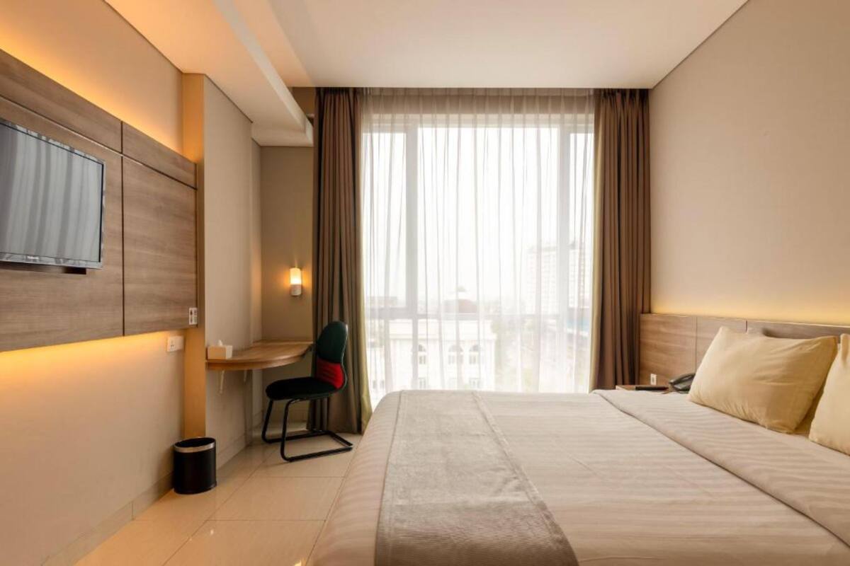 A hotel room with a bed, a desk, a chair, a TV mounted on the wall, and a window with curtains.