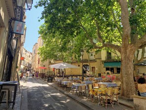 An outdoor café with tables and chairs, a tree providing shade, and a street with buildings on one side.