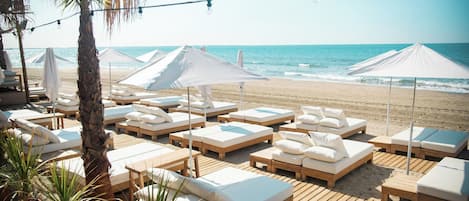 Un área de descanso frente a la playa con tumbonas de madera y cojines blancos, dispuestas sobre una terraza de madera con sombrillas que ofrecen sombra.
