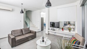 Un salon moderne avec un canapé en cuir marron, une table basse blanche et un téléviseur posé sur une console blanche. On aperçoit un escalier à l’arrière-plan.