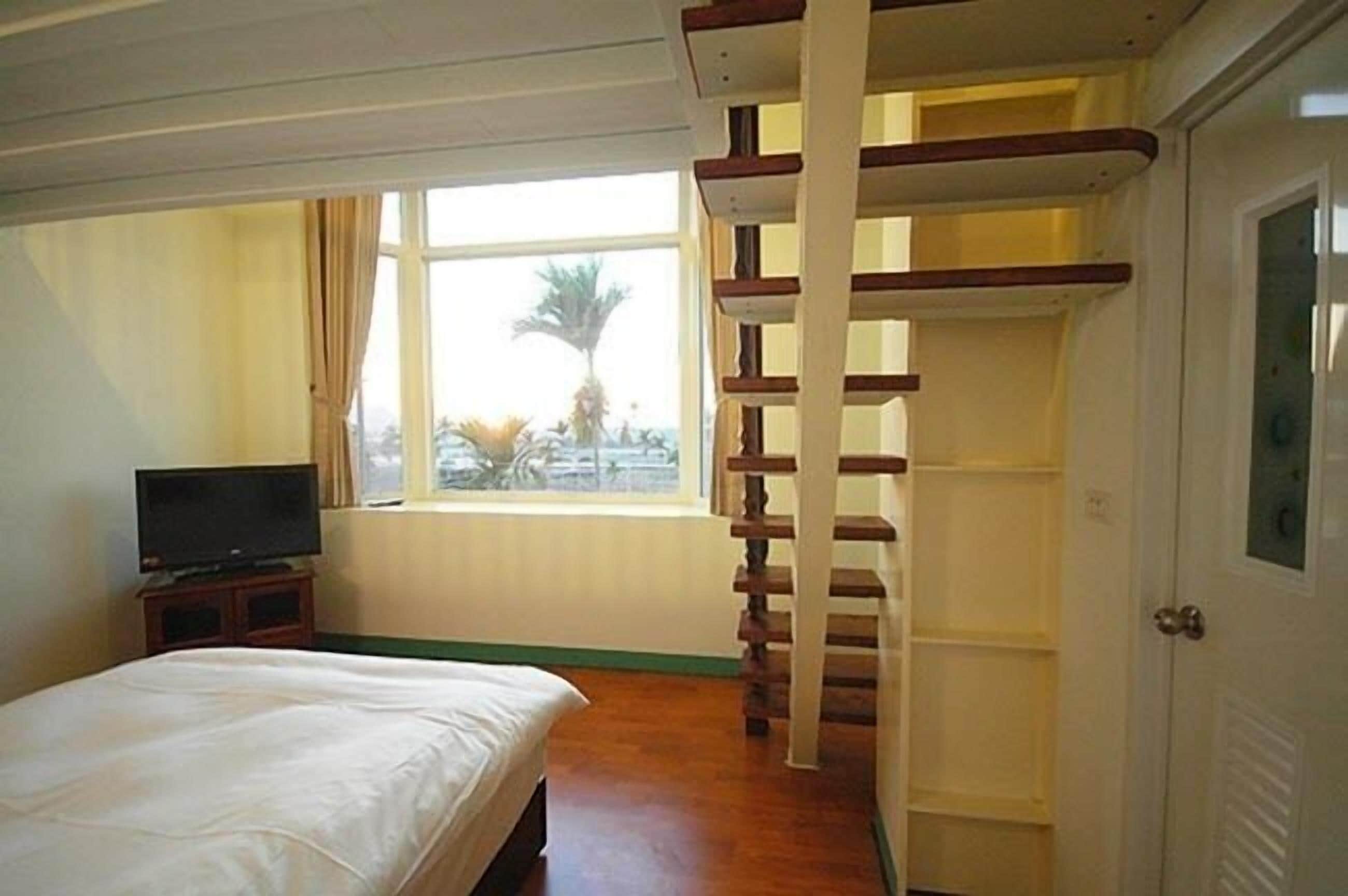 A bedroom with a bed, a TV on a stand, a window with a view of palm trees, and a wooden staircase.