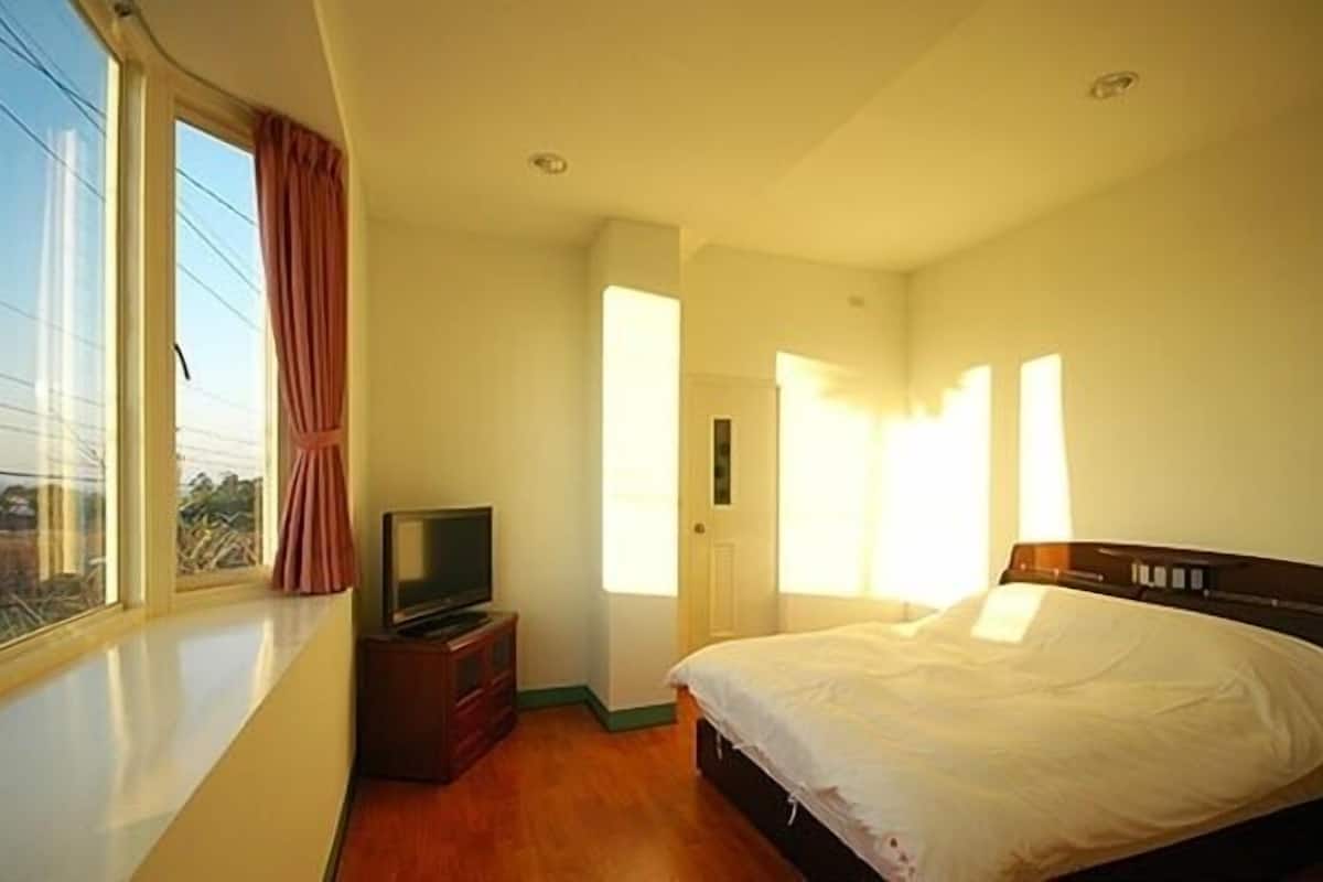 A hotel room with a large window, a bed, a TV on a wooden stand, and a door leading to another room.
