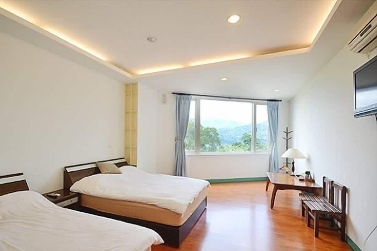 A hotel room with two beds, a desk, a chair, and a large window with a view of the mountains.
