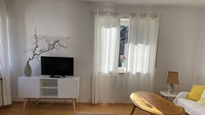 Un salon moderne avec un téléviseur à écran plat posé sur une console blanche, une table basse en bois, un canapé blanc agrémenté d’un coussin jaune et un tapis à motifs.