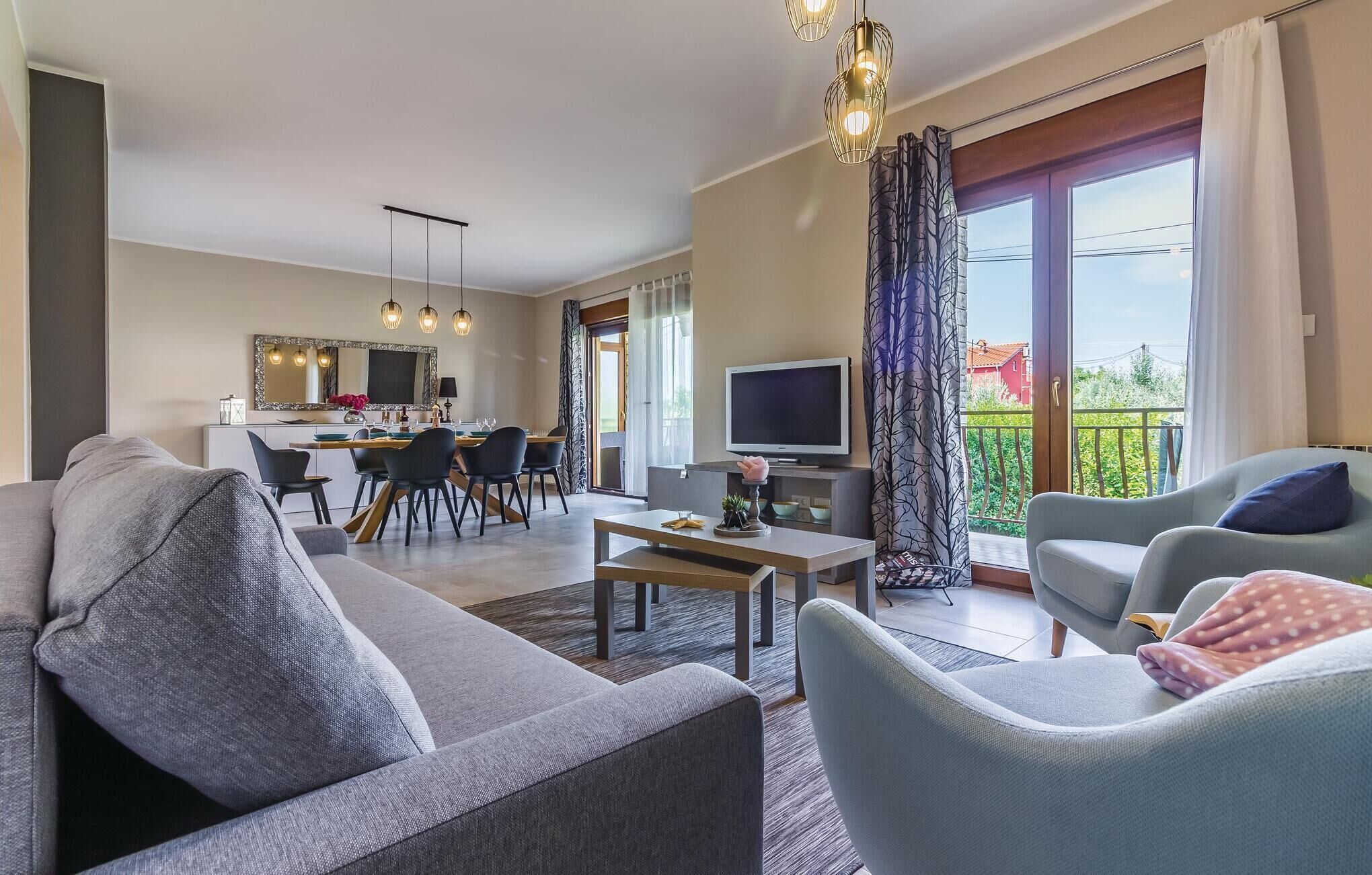 A modern living room with a grey sofa, a coffee table, and a TV. There is a dining area with a table and chairs, and a balcony with a view.