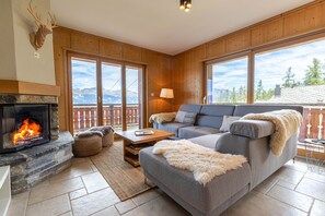Un salon chaleureux avec une cheminée, un canapé gris, une table basse en bois et une vue sur les montagnes par de grandes fenêtres.