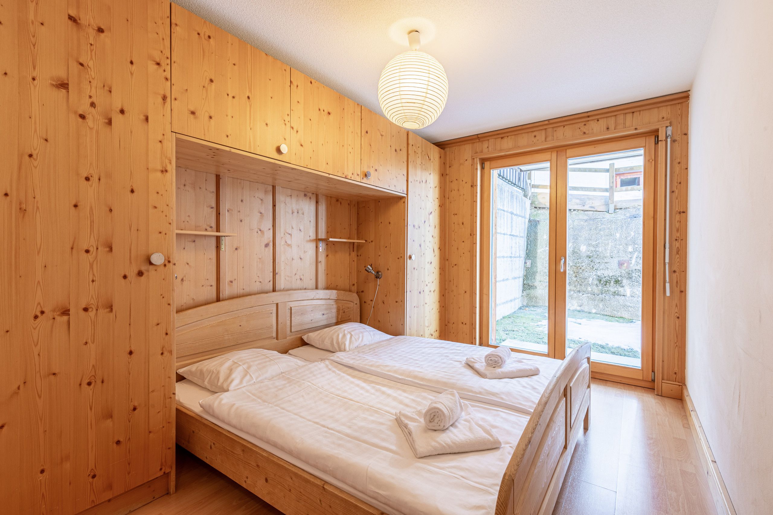Ein Schlafzimmer mit Holzboden, zwei Betten, einem großen Fenster und einer Hängeleuchte.