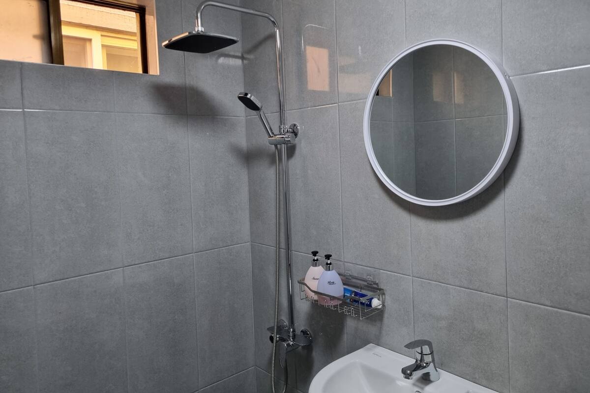 Un baño moderno con baldosas grises, ducha con cabezal de ducha manual, espejo redondo y un estante pequeño con artículos de aseo.