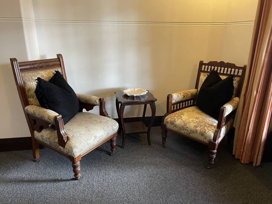 Two wooden armchairs with floral upholstery and black cushions, a small wooden side table with a white bowl, and a carpeted floor.