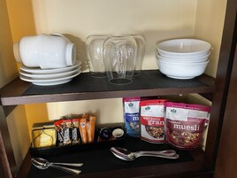 Una estantería con vajilla blanca, vasos transparentes y productos para el desayuno, incluyendo mezcla Pukka English Breakfast y Nescafé.