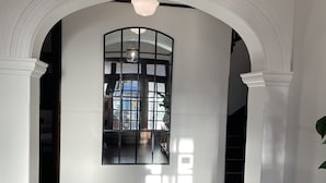 Een gang met een grote gewelfde deuropening naar een kamer met een groot raam. De vloer is van hout en er ligt een tapijt in de gang.