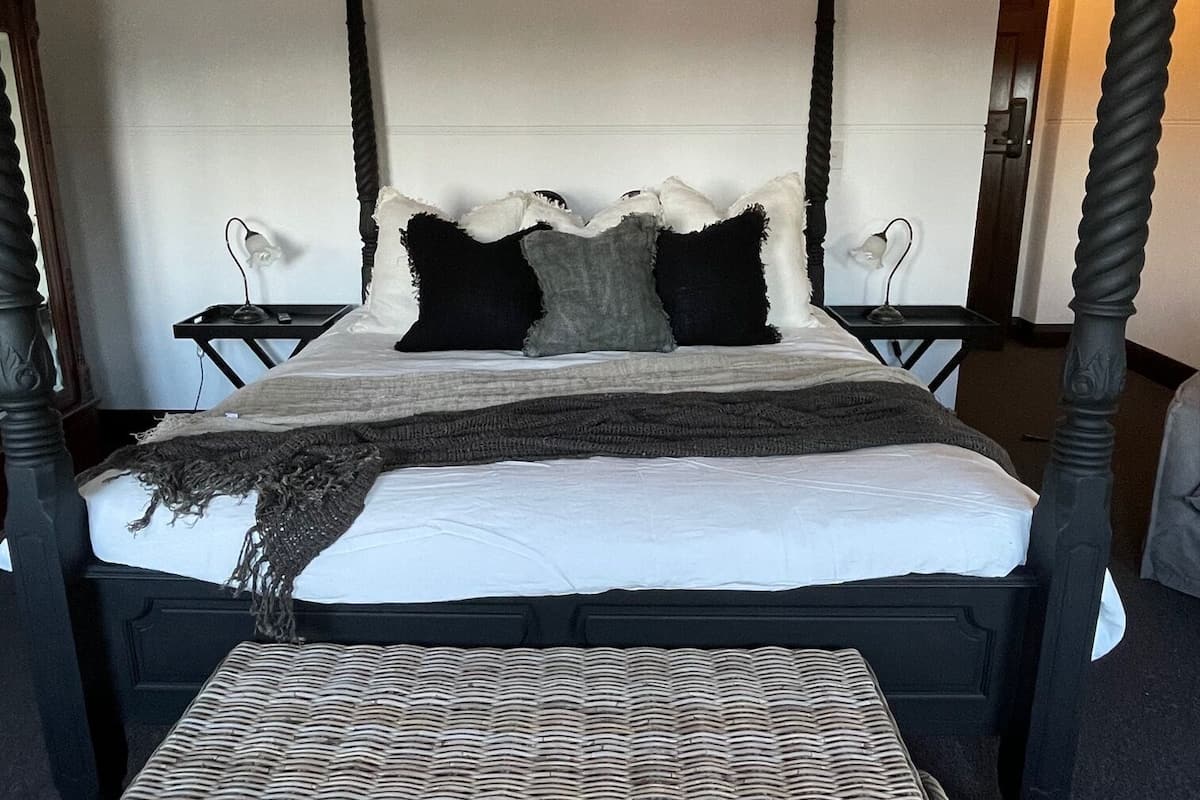 Ein Bett mit Himmel, dessen dunkles Holzgestell einen weißen Lattenrost und eine graublaue Decke trägt. Vor dem Bett steht ein Korbsitzhocker.