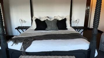 Una cama con dosel de madera oscura, un colchón blanco y una manta de color gris y negro. Hay un reposapiés de mimbre frente a la cama.
