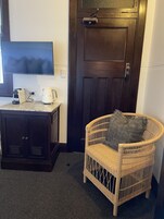 Una habitación con una puerta de madera oscura, una silla de mimbre con cojín, una mesita con cafetera y un televisor montado en la pared.
