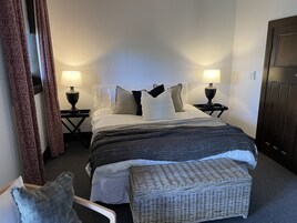 Un dormitorio ordenado con una cama, dos mesitas de noche con lámparas, un banco de mimbre y un armario.
