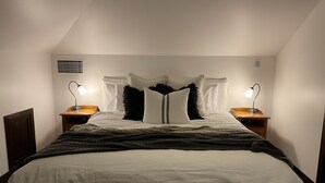 Een net opgemaakt bed met een witte deken en een donkere, gemêleerde plaid. Op de houten nachtkastjes staan twee bedlampjes.