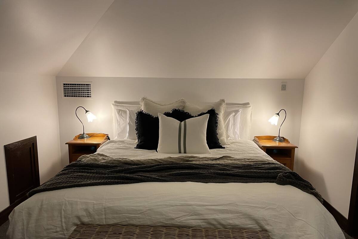 Ein ordentlich bezogenes Bett mit weißer Decke und farbigem Überwurf. Auf Holznachttischen stehen je eine Nachttischlampe.