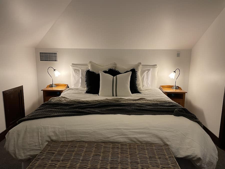 Ein ordentlich bezogenes Bett mit weißer Decke und farbigem Überwurf. Auf Holznachttischen stehen je eine Nachttischlampe.