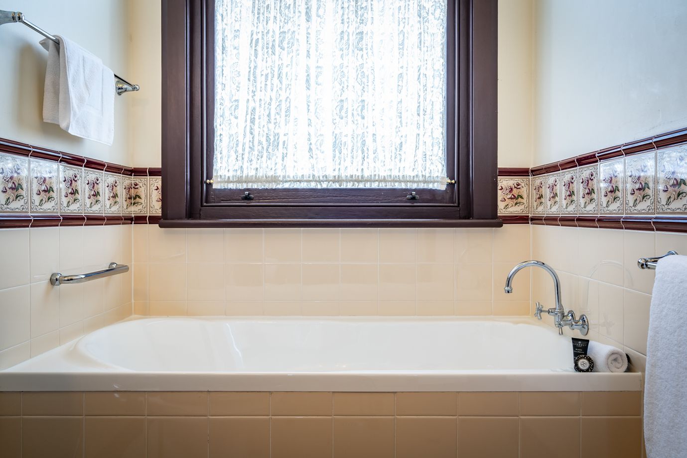 A bathroom with a bathtub, a window with lace curtains, and decorative tiles.