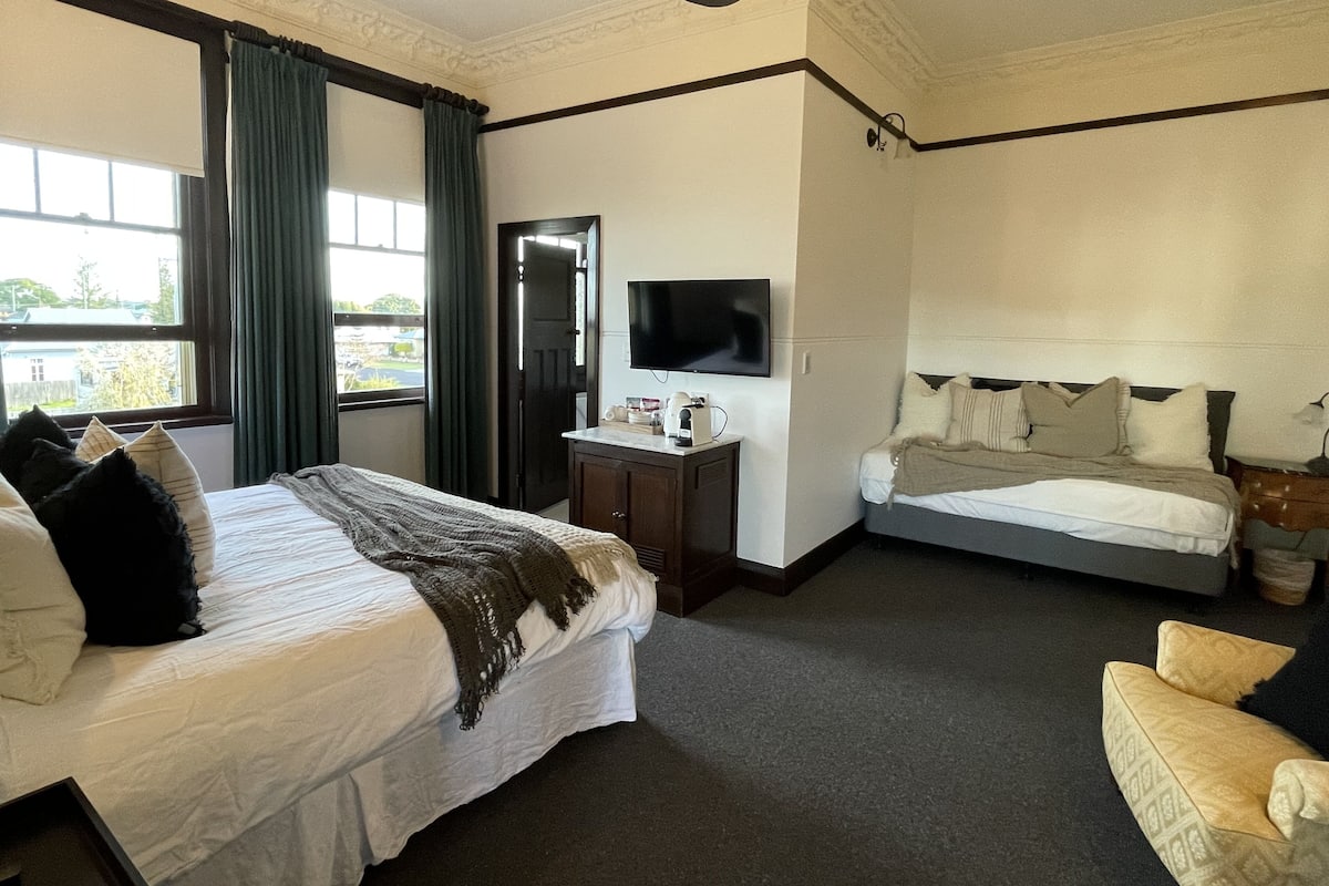 Ein Hotelzimmer mit zwei Betten, einem Schreibtisch, einem Flachbildfernseher, einem Deckenventilator und einem fenster mit Vorhängen.
