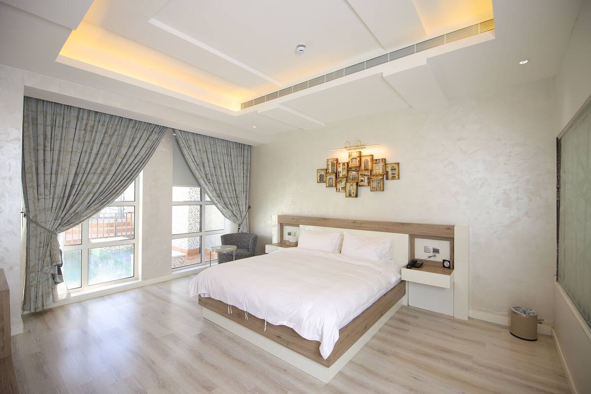 A bedroom with a large bed, a bedside table, a wall-mounted light fixture, and a view through a window with curtains.