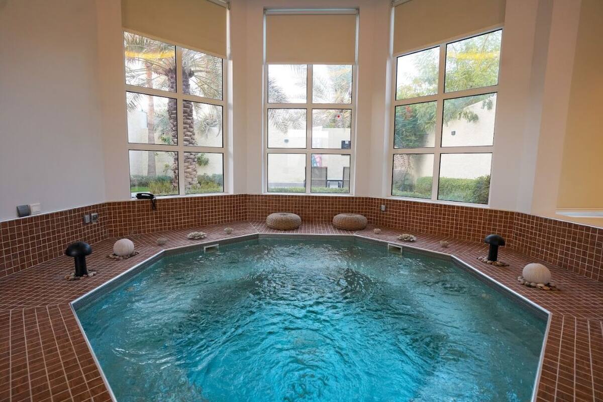 A large indoor swimming pool with a tiled border, large windows, and decorative stones.