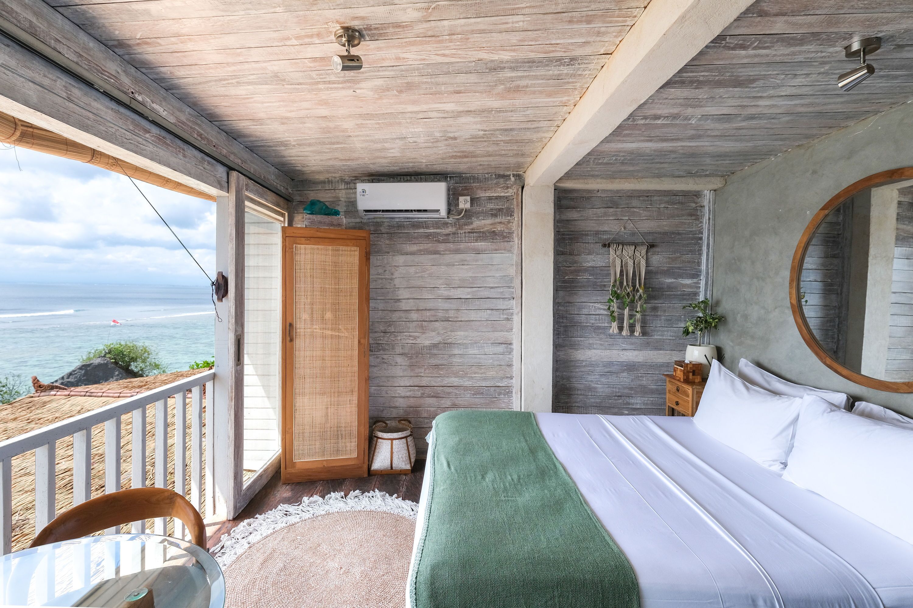 A bedroom with a large bed, a wooden ceiling, a view of the sea, a small table, and a nightstand with a lamp.