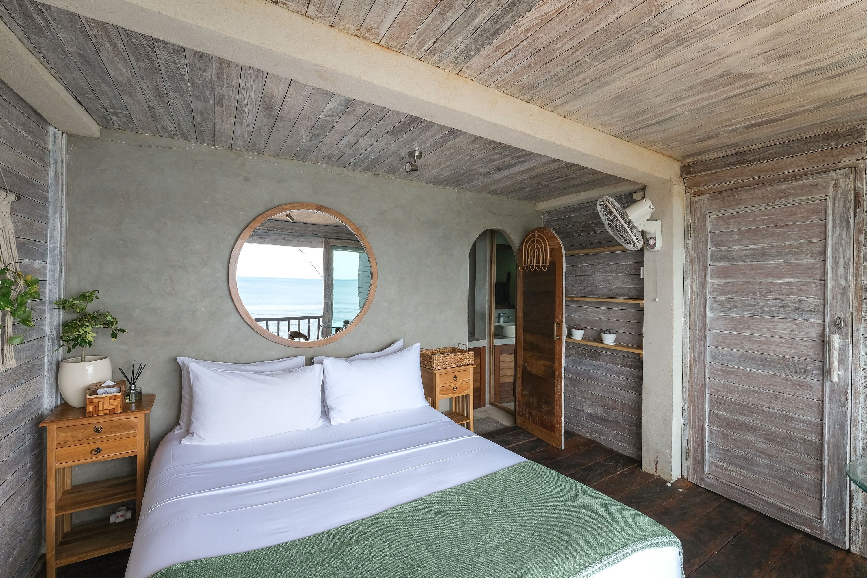 A bedroom with a wooden ceiling, a large bed with white bedding, a round mirror, a wooden wardrobe, and a view of the sea.