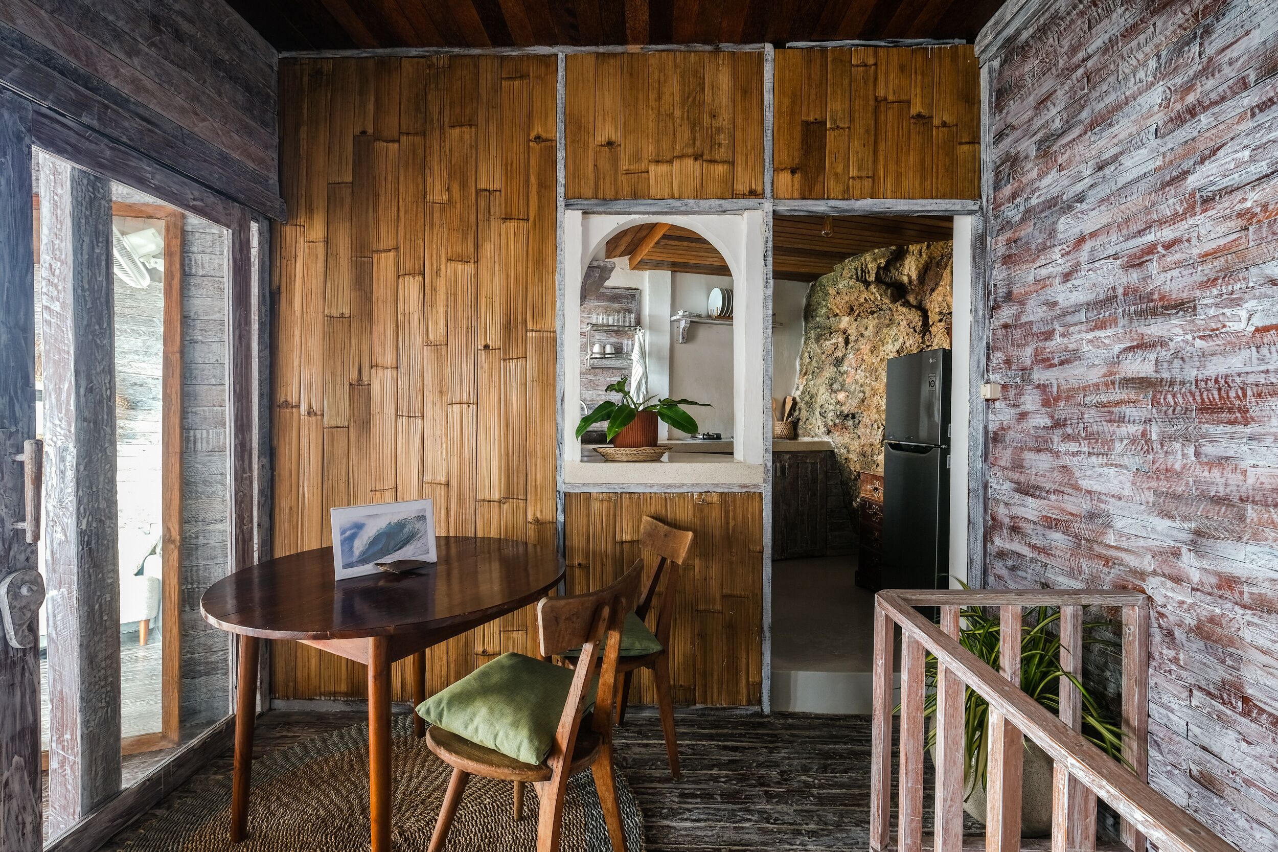 A dining area with a wooden table and chairs, a small kitchen visible through an archway, and a rustic brick wall.