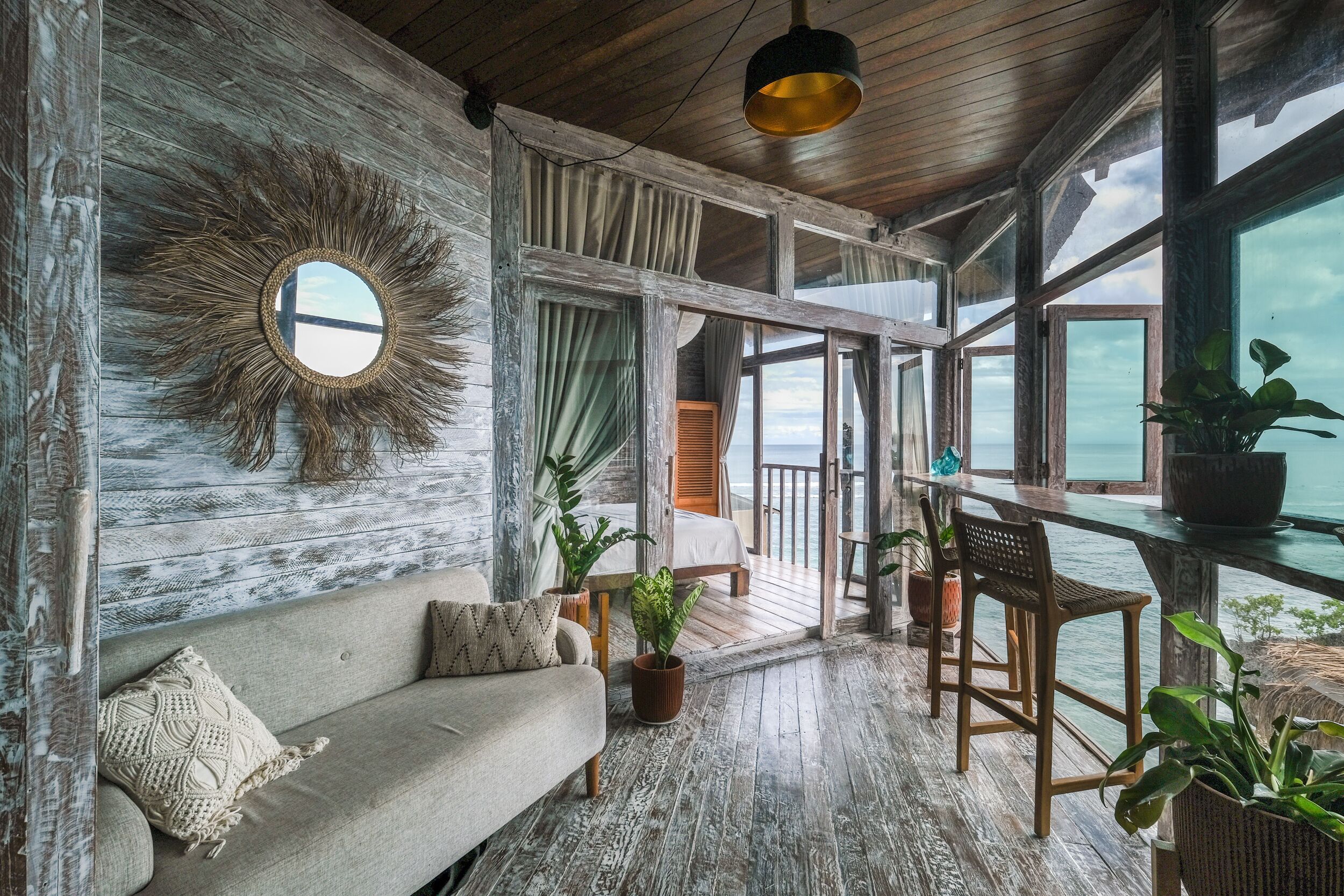 A cozy room with a wooden ceiling, a round mirror, a sofa, and a view of the sea through large windows.