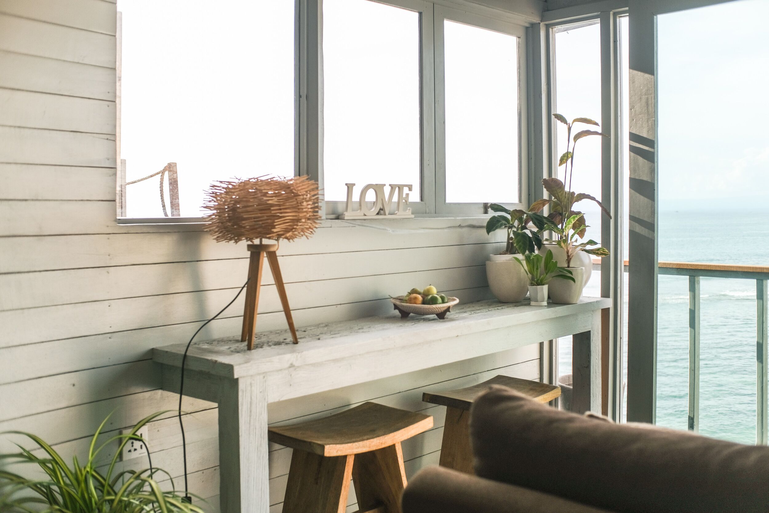Una habitación con un ventanal grande con vista al mar, una consola de madera con un espejo, una lámpara de mimbre y un cuenco de fruta.