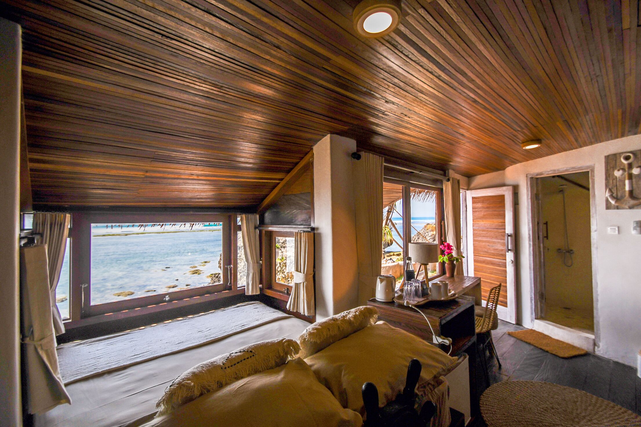 A room with a wooden ceiling, a bed with white bedding, a desk with a lamp, and a view of the sea through large windows.