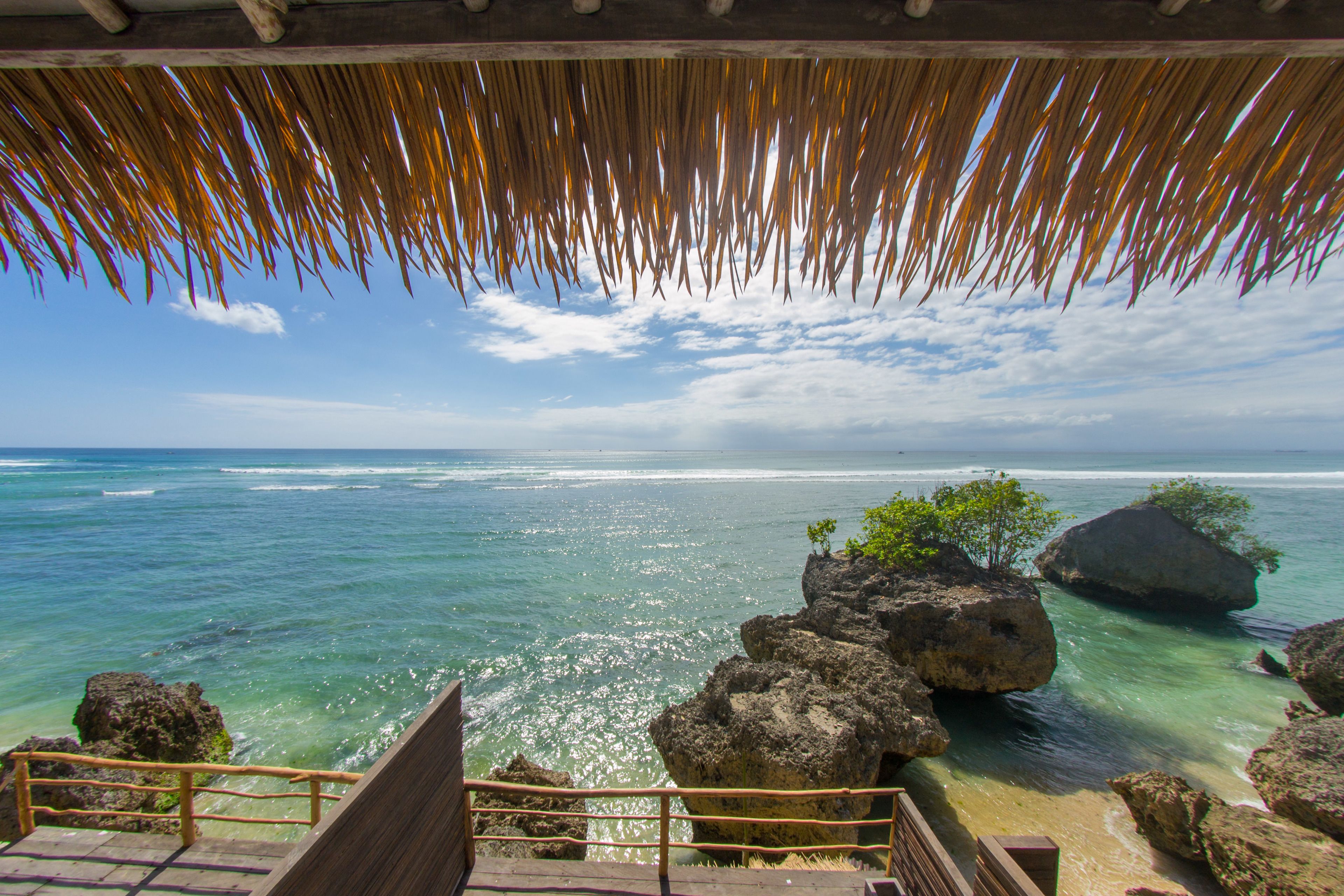 A wooden staircase leading to a beach with clear turquoise water, large rocks, and a thatched roof structure.