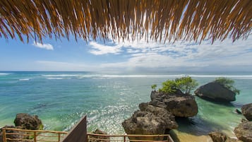 A wooden staircase leading to a beach with clear turquoise water, large rocks, and a thatched roof structure.
