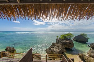 A wooden staircase leading to a beach with clear turquoise water, large rocks, and a thatched roof structure.
