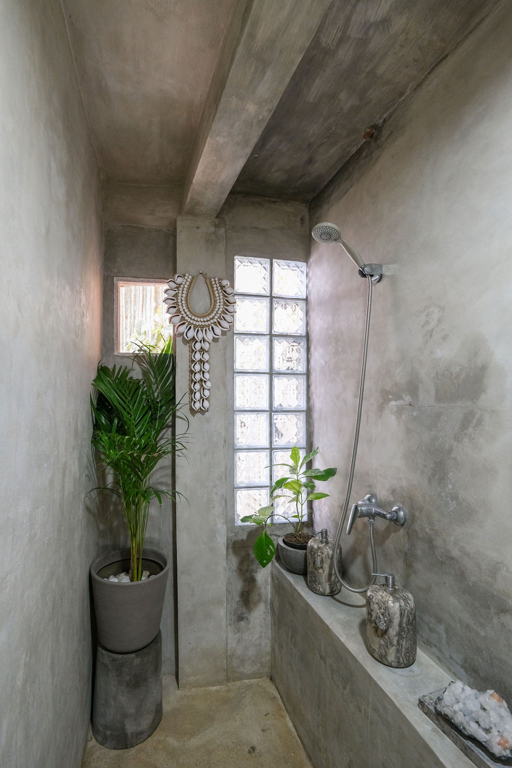 Ein kleines, schmales Badezimmer mit Betonfussboden und -wänden, einem Fenster mit dekorativem Spiegel und einem Duschbereich inklusive Handbrause.